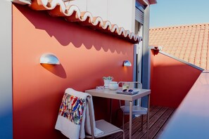 Un balcón con una pared roja, una mesa blanca, una silla, una toalla, una planta en maceta y una lámpara de mesa pequeña.