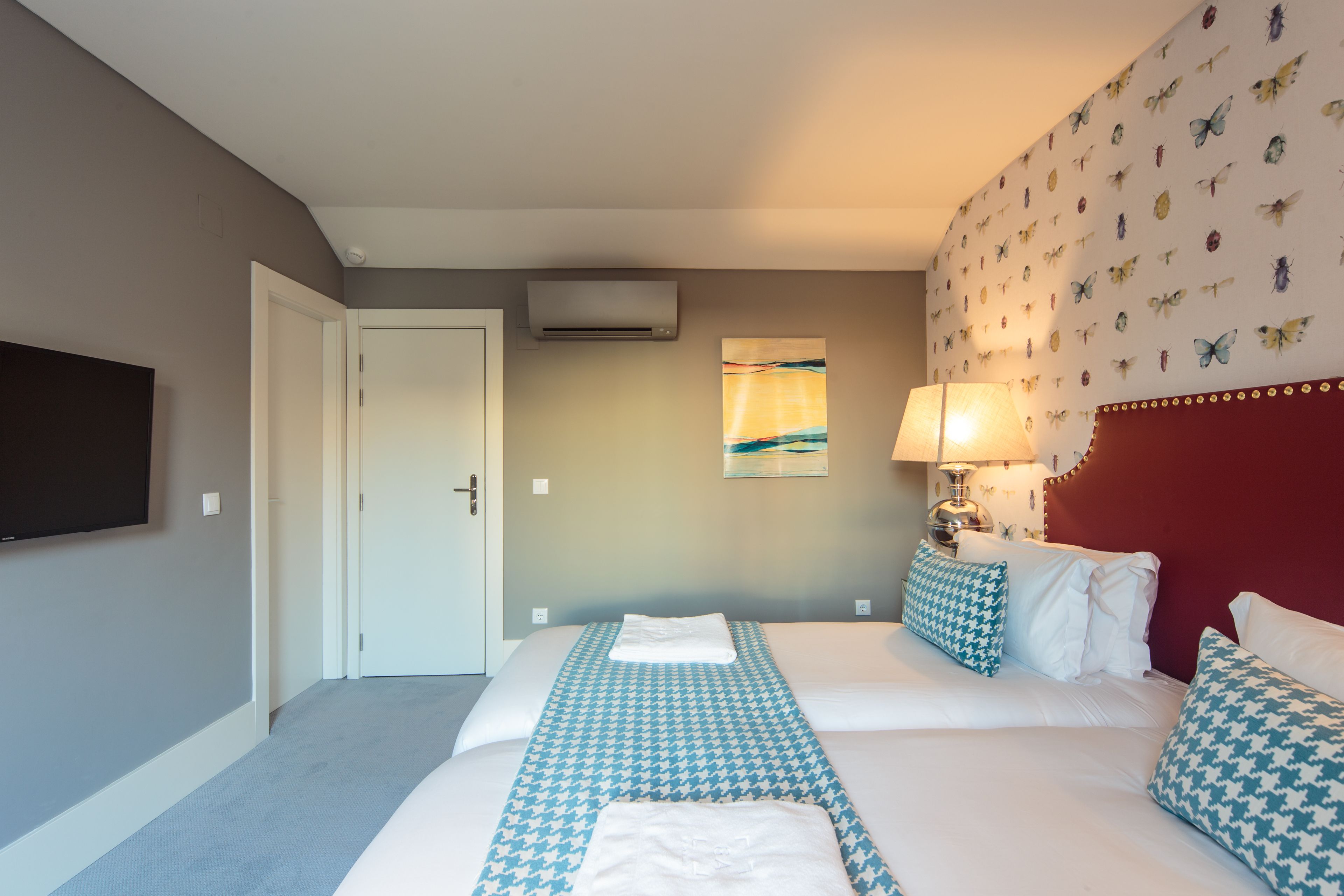 Una habitación de hotel con una cama, televisión, aire acondicionado montado en la pared, una puerta y un papel tapiz estampado.