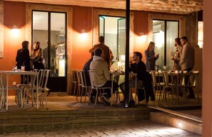 Un área de comedor al aire libre con mesas y sillas, una terraza de madera y una entrada con puerta de vidrio.