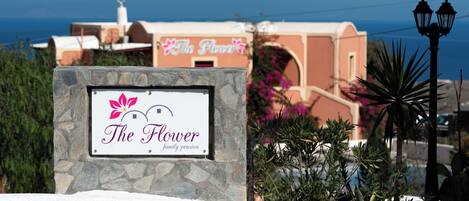 A sign for 'The Flower Family Pension' with a stone frame, a view of a building with a pink facade, and a clear blue sky.