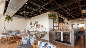 A modern interior with wooden flooring, a seating area with cushioned chairs, a bar area with stools, and hanging plants.