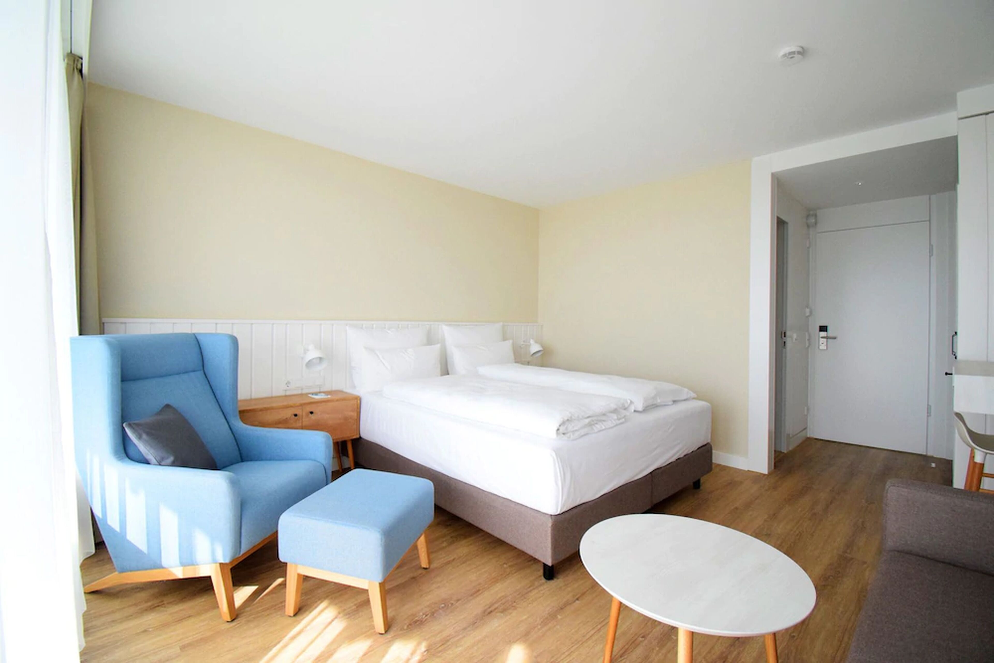 Een moderne hotelkamer met een groot bed, een blauwe fauteuil, een klein voetenbankje en een ronde tafel.