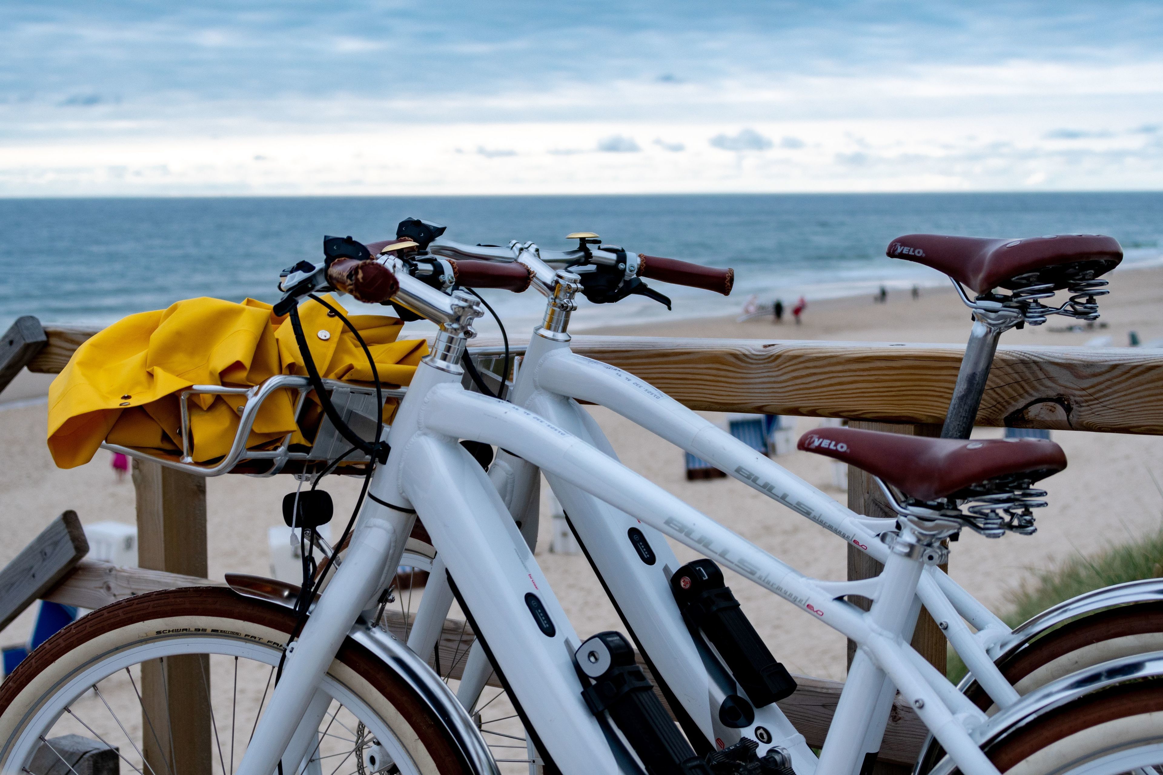 Una bicicleta blanca con una bolsa amarilla y un asiento rojo estacionada sobre una cubierta de madera junto a la playa.