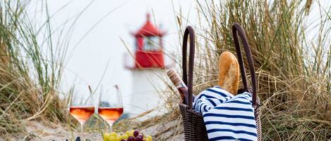 A wicker basket with a striped towel and baguette, a plate of grapes, and two glasses of red wine on a blanket, with tall grass and a red lighthouse in the background.