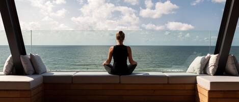 A person sitting on a wooden bench with cushions, facing the ocean.