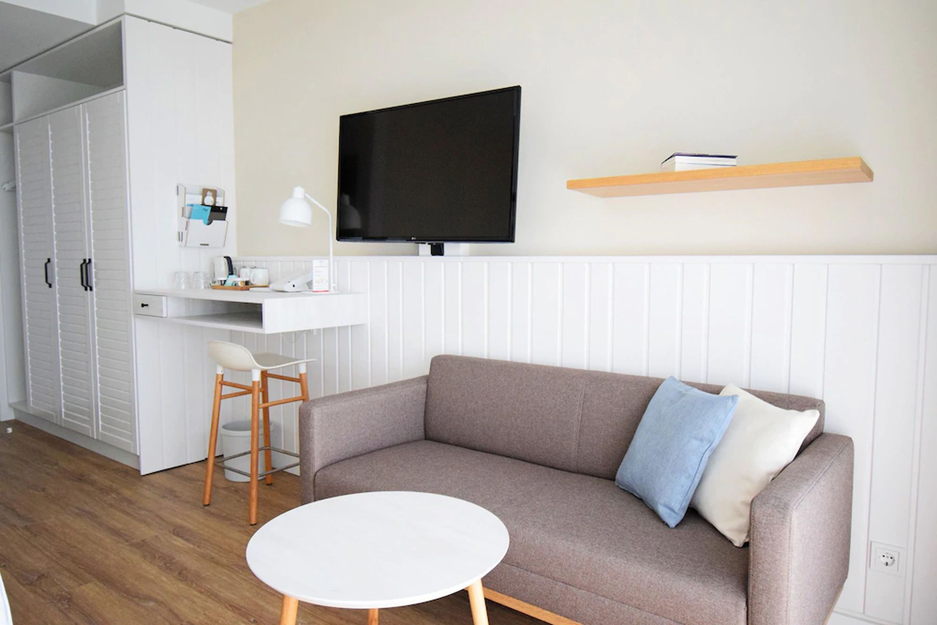 A modern living room with a sofa, a round table, a TV mounted on the wall, and a small shelf with books.