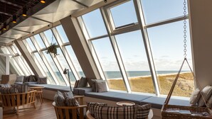 A room with large windows overlooking a beach, furnished with wooden chairs and a sofa with plaid cushions.