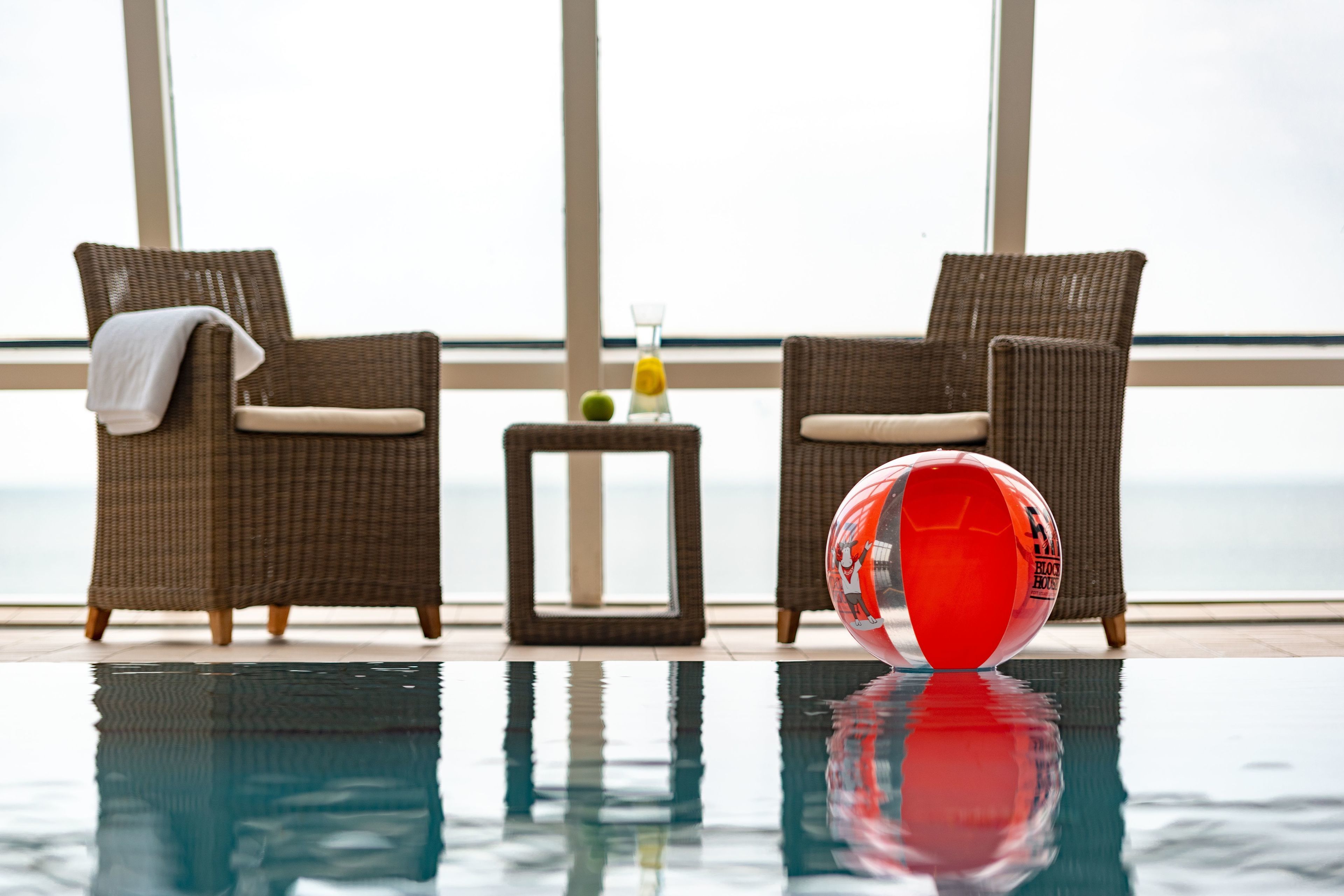 A poolside area with two wicker chairs, a small table with a glass and a bottle, and a red and white beach ball.