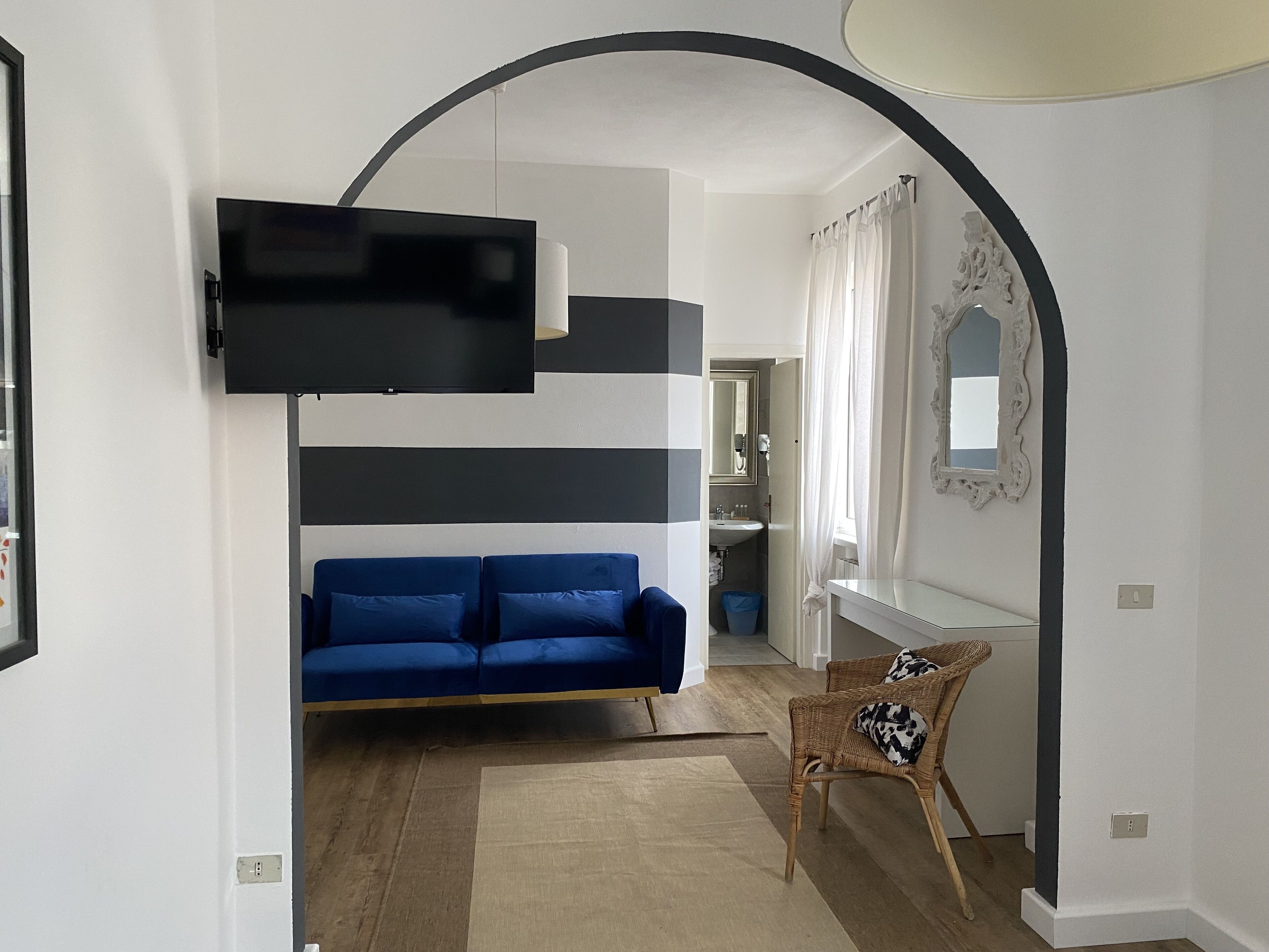 A modern living room with a blue sofa, a flat-screen TV mounted on a black frame, a wicker chair, and a white wall with a decorative mirror.
