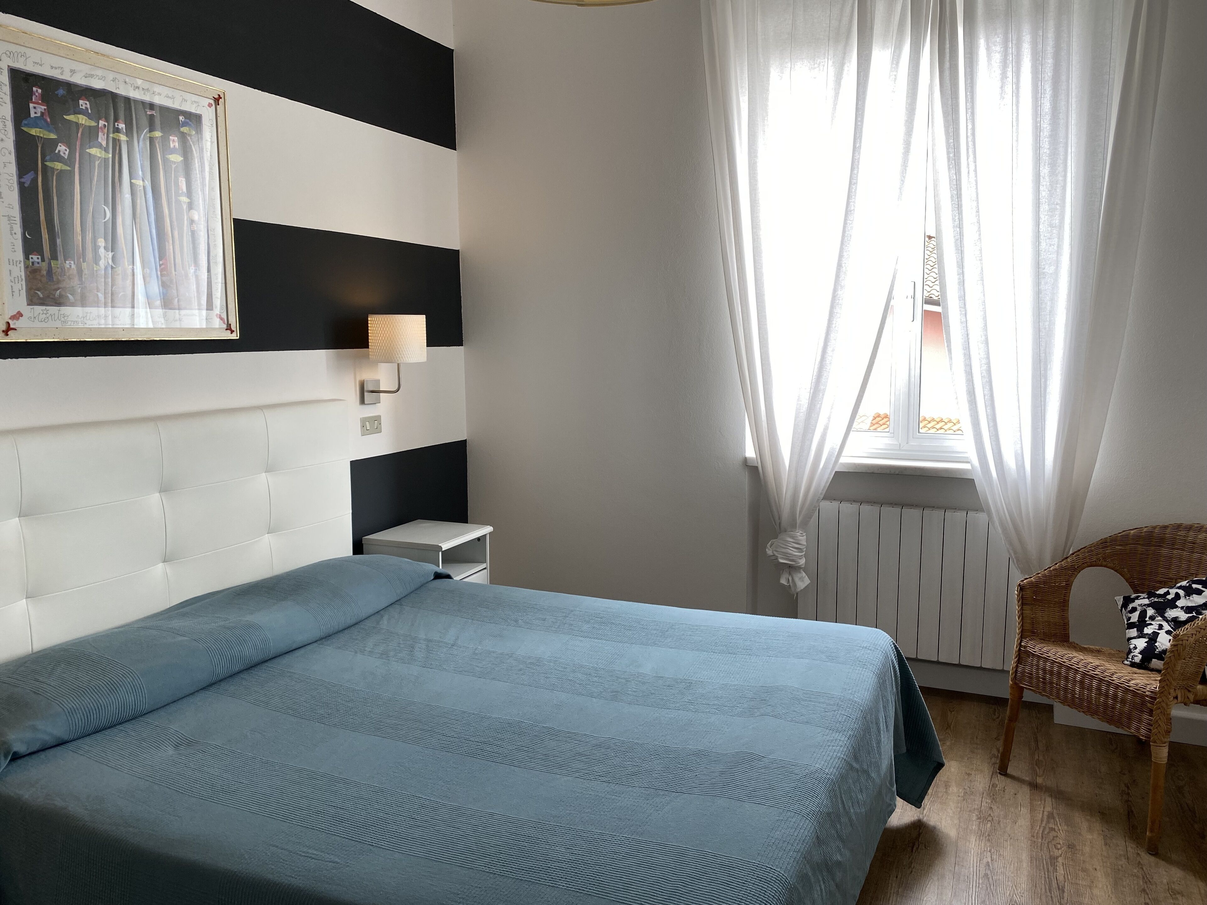 Un dormitorio con cama, una mesita de noche, una lámpara de pared, una silla de mimbre y una ventana con cortinas traslúcidas.
