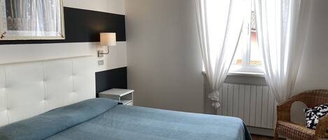 Un dormitorio con cama, una mesita de noche, una lámpara de pared, una silla de mimbre y una ventana con cortinas traslúcidas.