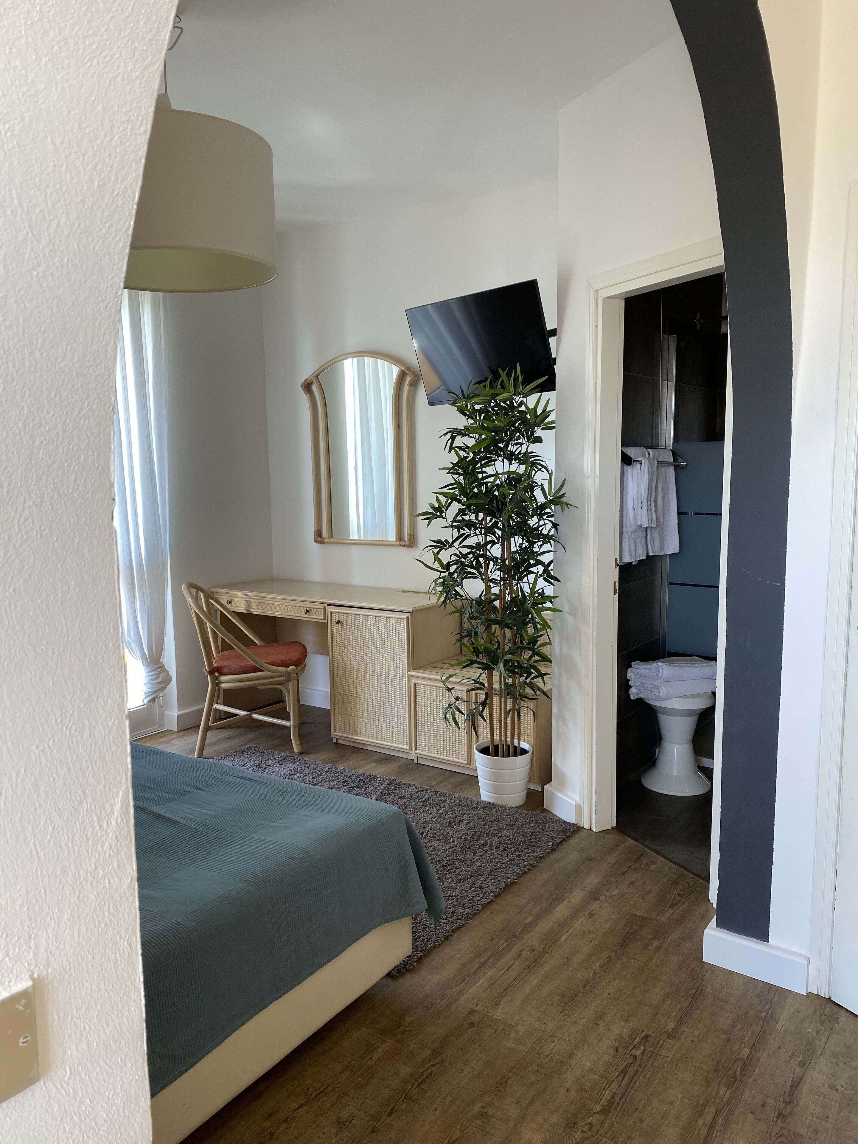 Un dormitorio con cama, escritorio, silla, planta en maceta y baño visible a través de un arco.