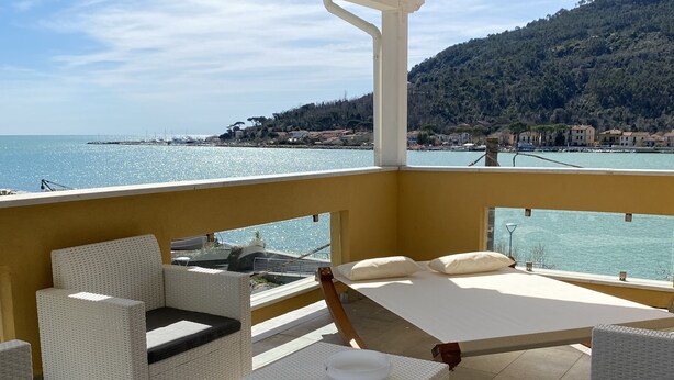 Een balkon met rieten meubilair, een bed en uitzicht op zee en heuvels.