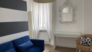 Ein Zimmer mit einem blauen Sofa, einem weißen Konsolentisch, einem Korbstuhl, einem großen Spiegel und einem Fenster mit transparenten Vorhängen.
