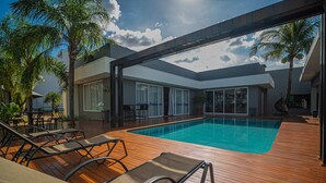 Zona de piscina con sillones de descanso, palmeras y un edificio moderno con ventanales grandes.