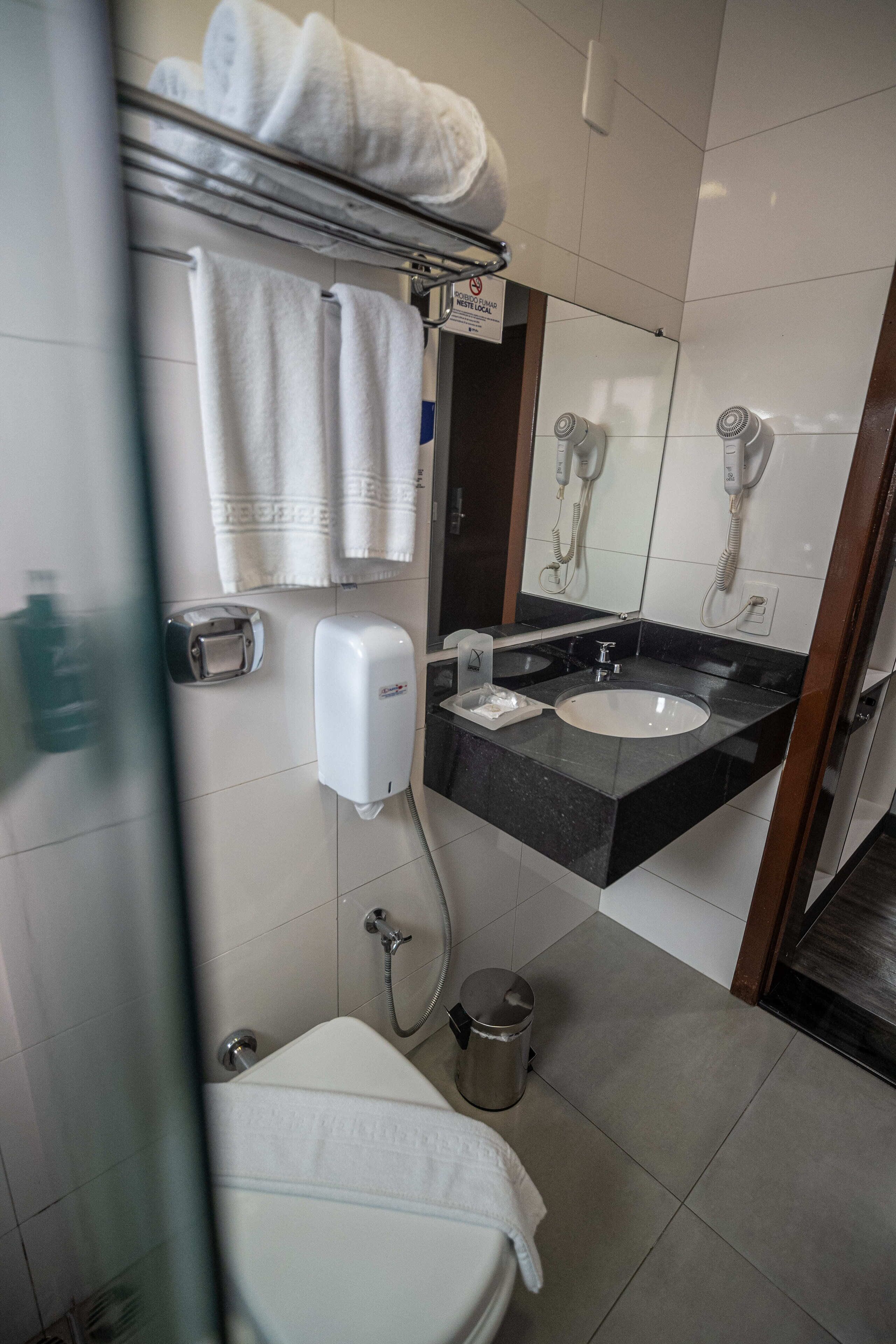 A bathroom with a toilet, sink, towel rack, hand dryer, and a trash can.
