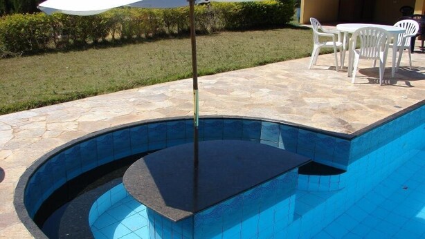 A swimming pool with a curved edge, a stone patio, and a white umbrella. - Chácara Marson (Mandaguaçu)