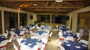 A banquet hall with multiple round tables set with white chairs and blue tablecloths, wooden beams on the ceiling, and large windows on the walls. - Chácara Marson (Mandaguaçu)