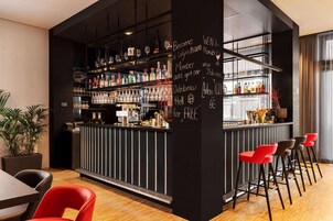 Zona de bar con estantes llenos de botellas y vasos, una pizarra con texto y taburetes rojos.
