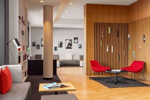 A modern lobby with a sofa, red chairs, a table with a book, a bookshelf, and a wooden wall with pictures. - Ernst Leitz Hotel Wetzlar (Wetzlar)