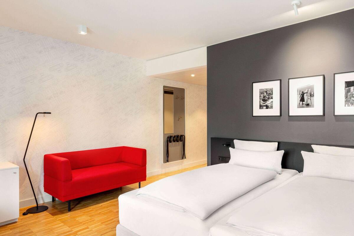 A modern hotel room with a large bed, a red sofa, a floor lamp, and artwork on the wall.