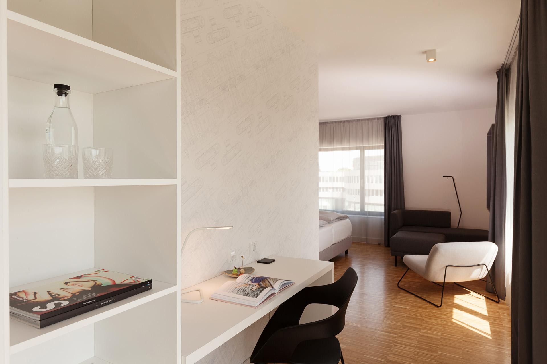 A modern hotel room with a white desk, a black chair, a sofa, and a large window with curtains.