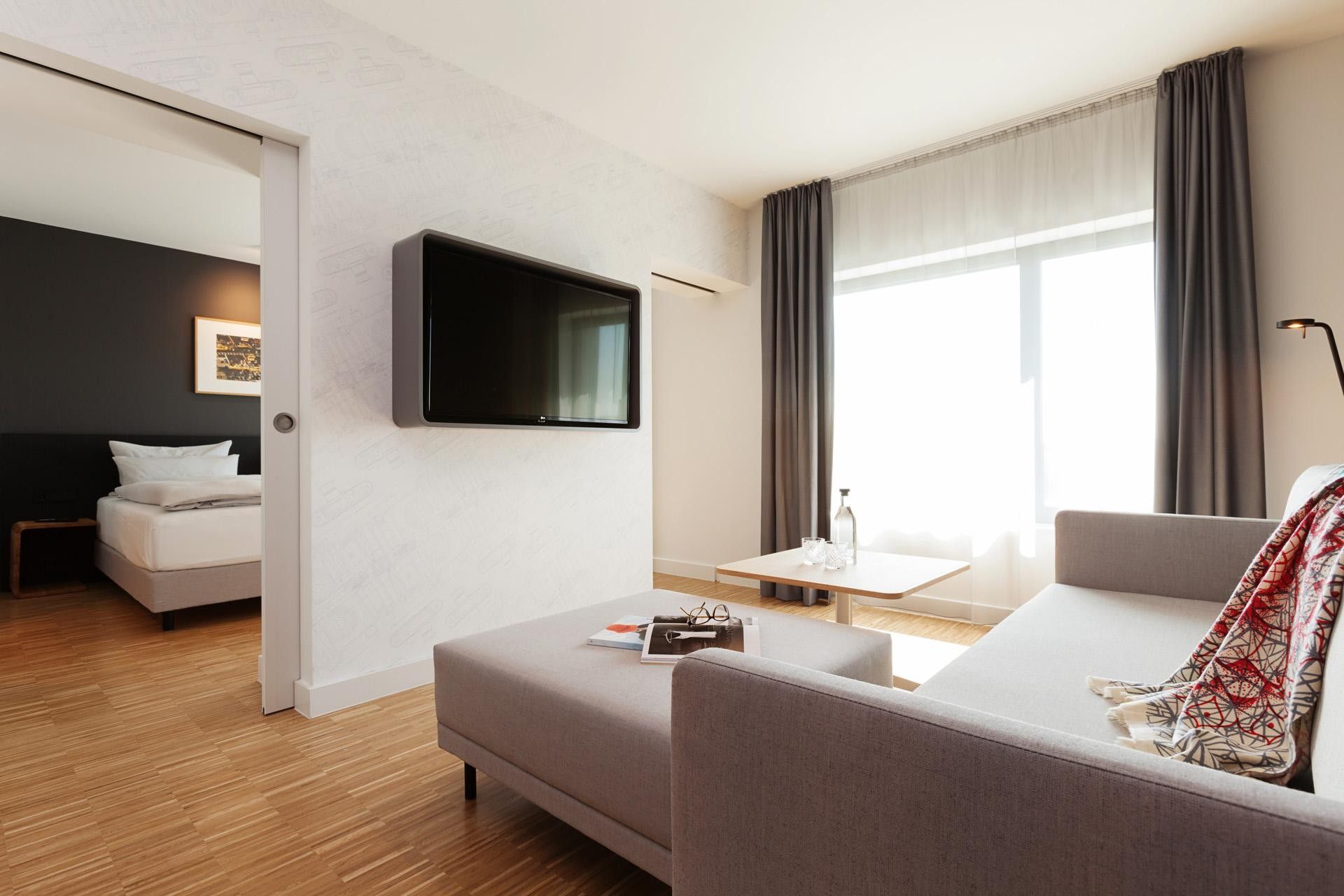 A modern hotel room with a flat-screen TV mounted on the wall, a sofa, a small table with glasses and a bottle, and a bed with white bedding.