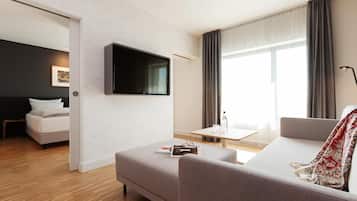 A modern hotel room with a flat-screen TV mounted on the wall, a sofa, a small table with glasses and a bottle, and a bed with white bedding.