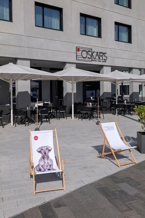 An outdoor seating area with tables and chairs, a building with a sign reading 'OSKARS,' and two easels with posters. - Ernst Leitz Hotel Wetzlar (Wetzlar)