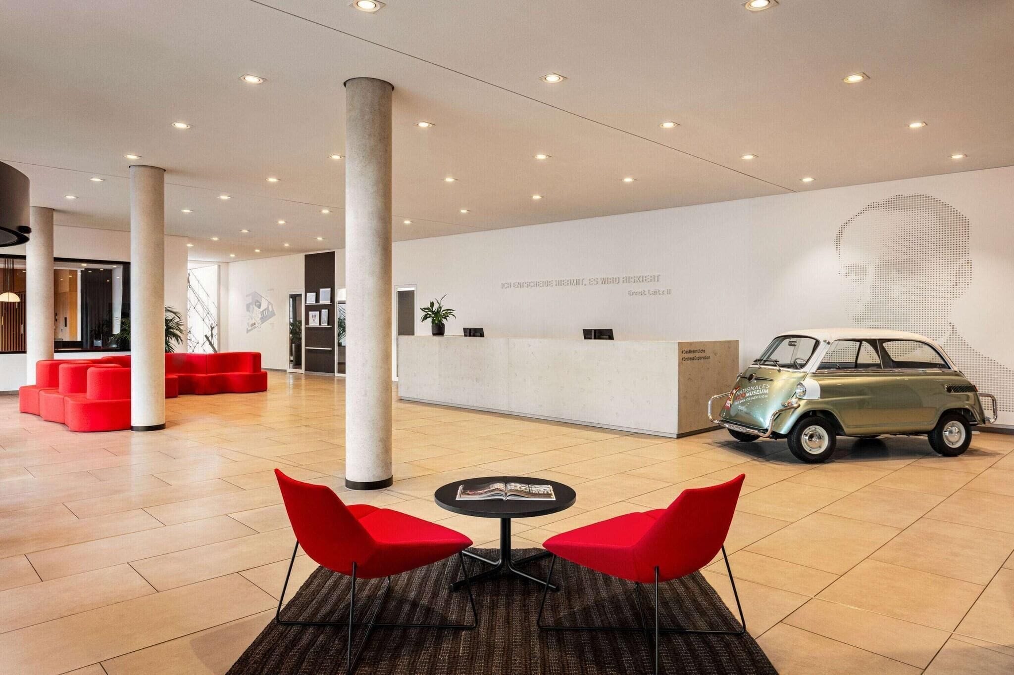 Un hall d’entrée moderne avec un comptoir d’accueil, une voiture ancienne, des sièges rouges et une grande colonne.