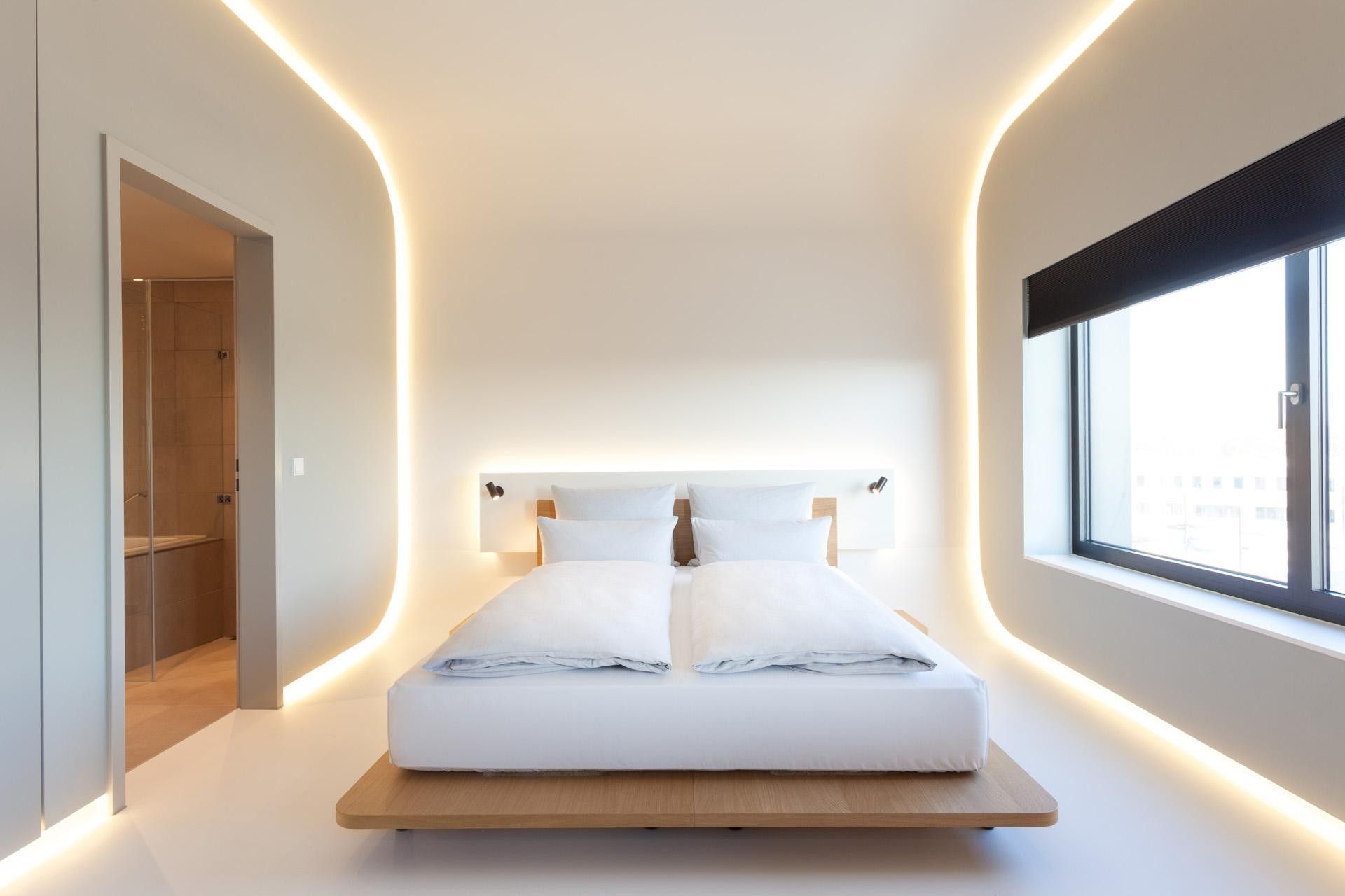 A modern bedroom with a bed centered on a platform, a large window on one side, and a shower area on the other.