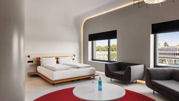 Una habitación de hotel moderna con una cama, dos sofás, una mesa de centro redonda y grandes ventanales con vistas a la ciudad.