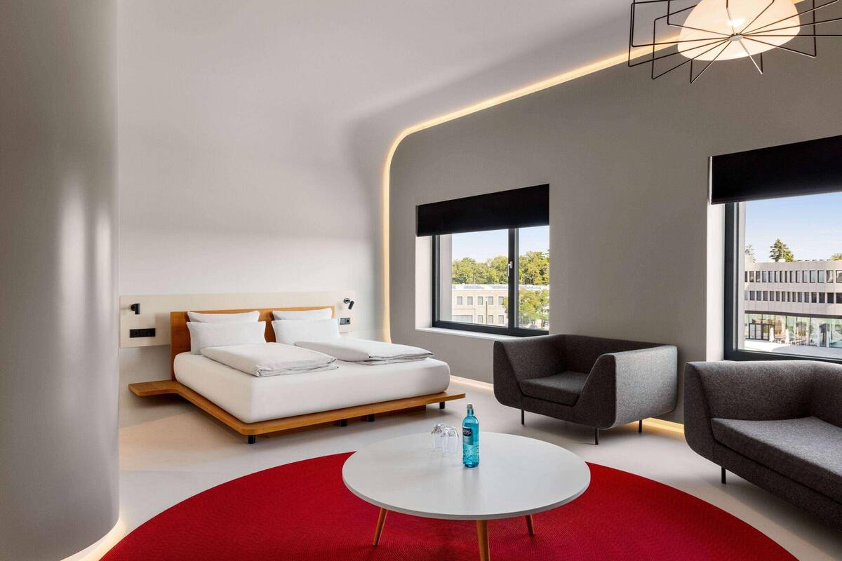 A modern hotel room with a bed, two sofas, a round coffee table, and large windows with a city view.