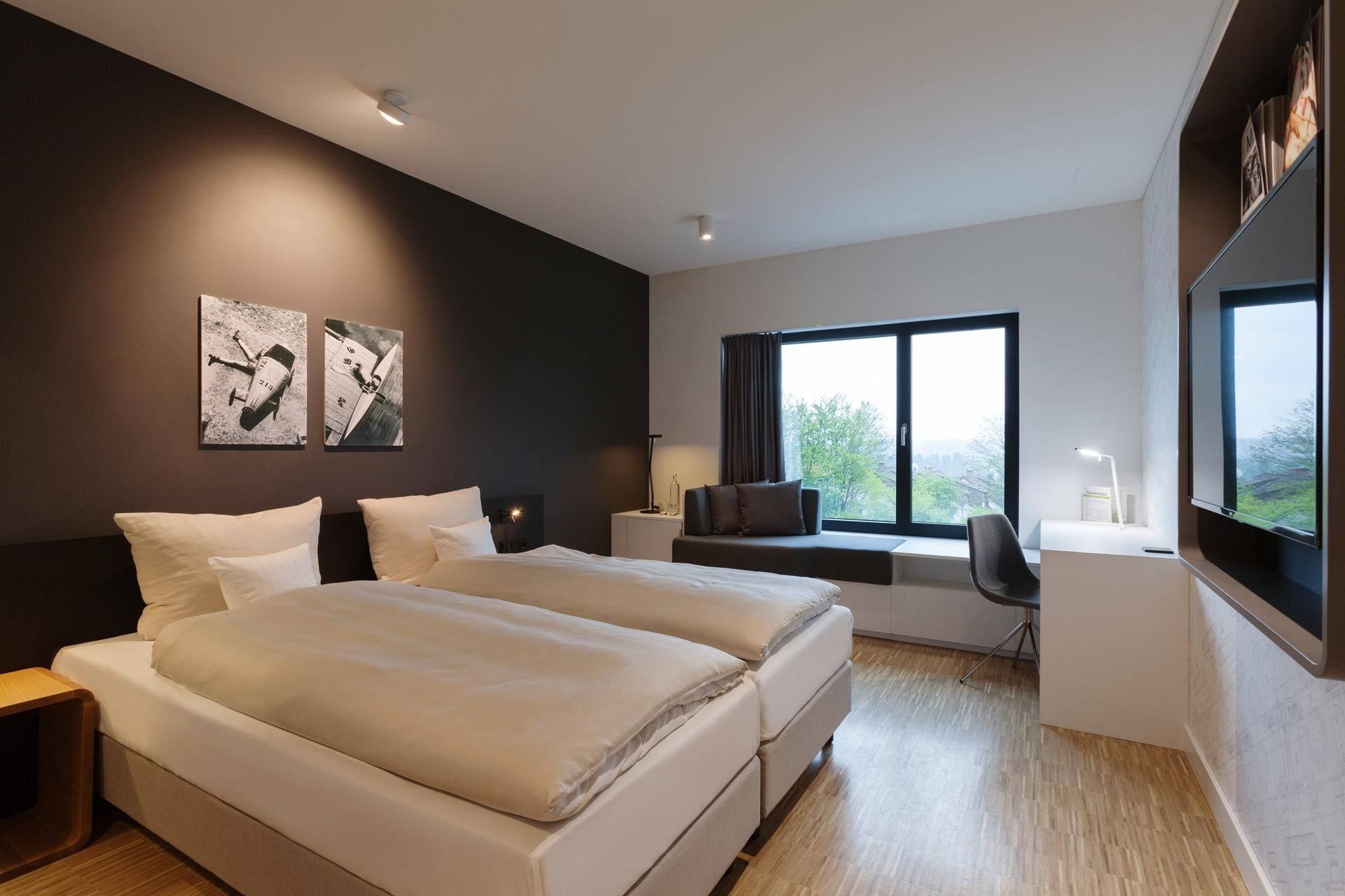 Ein modernes Schlafzimmer mit einem großen Bett, einem Schreibtisch und einem Stuhl. Zwei Wandbilder und ein Fenster mit Blick ins Freie.