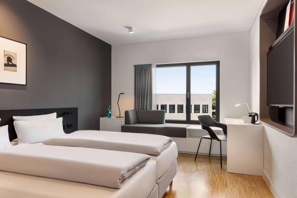 A modern hotel room with a bed, a desk, a chair, and a large window.