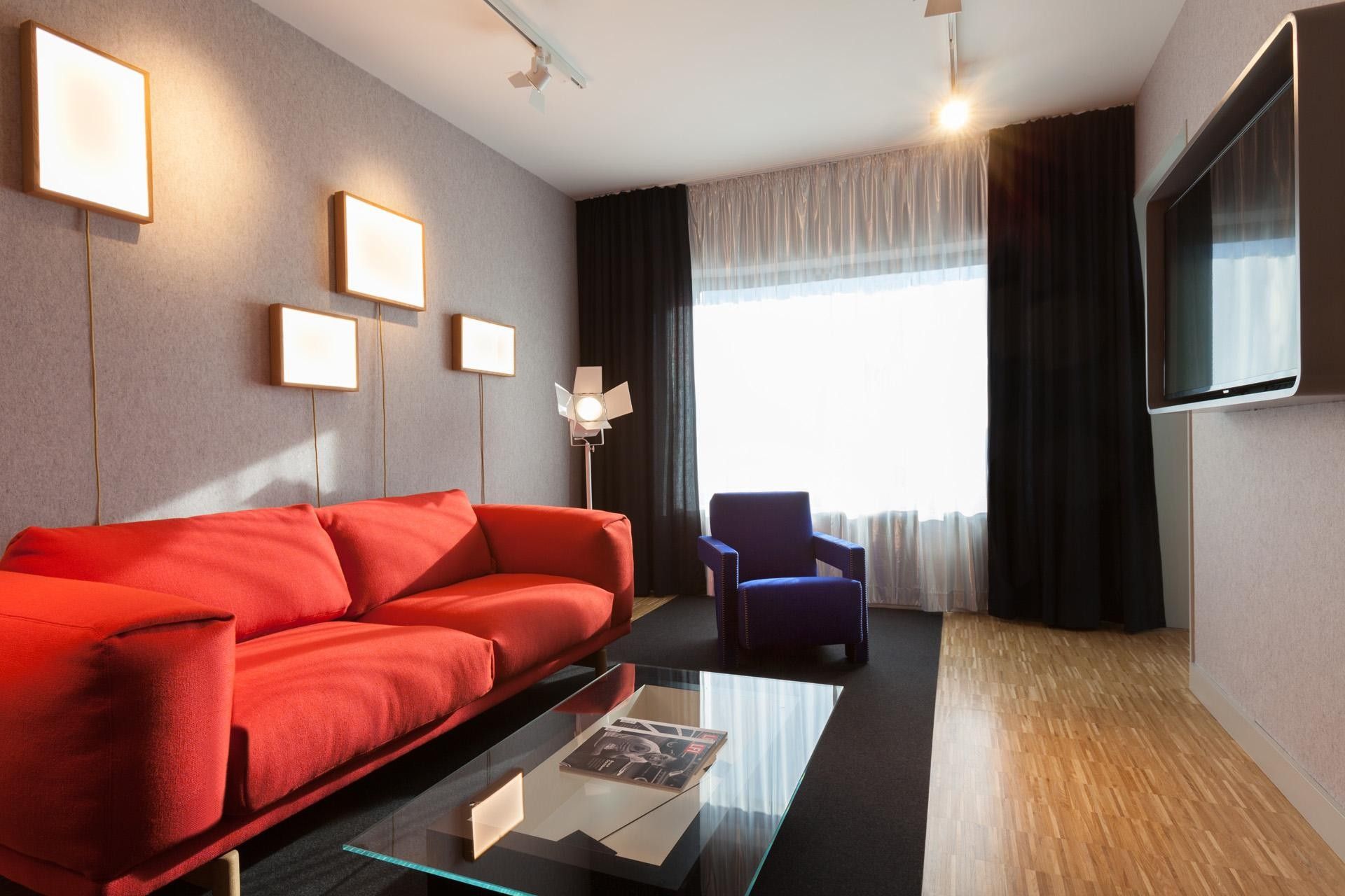 Een moderne woonkamer met een rode bank, een blauwe fauteuil, een glazen salontafel en een flatscreen televisie aan de muur.