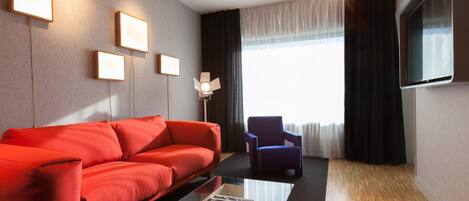 Een moderne woonkamer met een rode bank, een blauwe fauteuil, een glazen salontafel en een flatscreen televisie aan de muur.