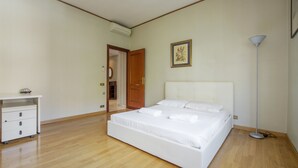Quarto com piso de madeira, uma cama branca, uma mesinha de cabeceira e um abajur.