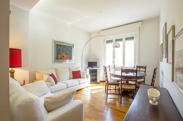 A living room with a white sofa, a dining table with chairs, a TV on a stand, and a painting on the wall.