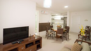 A living room with a flat-screen TV, a wooden entertainment unit, a dining area with a table and chairs, and a kitchen in the background. - Praia dos Ingleses Belo Recanto da Ilha (Florianópolis)