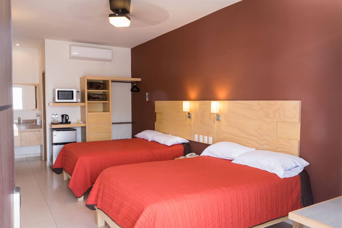 A hotel room with two beds, a kitchenette, and a wooden headboard with wall-mounted lights.