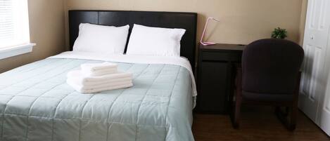 A neatly made bed with a light blue quilt and white pillows, a small desk with a chair, and a window with blinds.