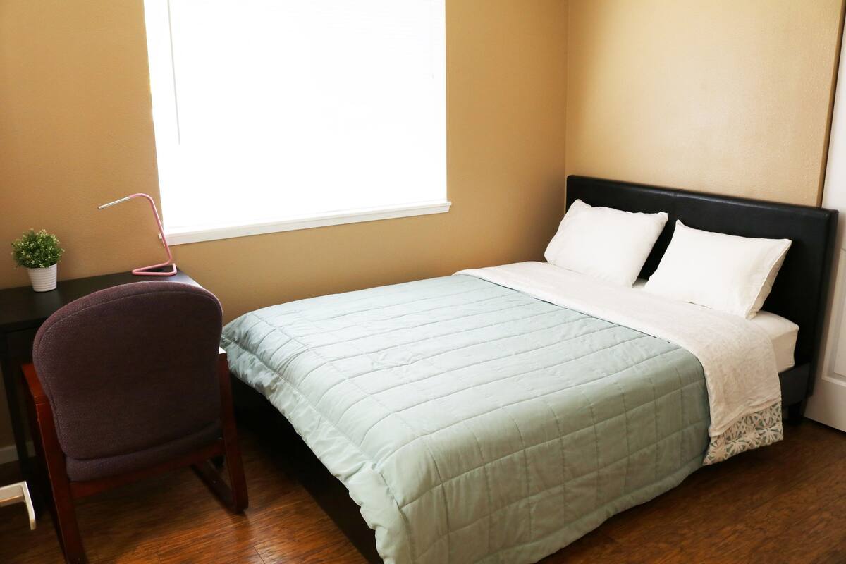 Ein Schlafzimmer mit einem Bett, einem Sessel, einem kleinen Tisch mit einer Pflanze und einem Fenster.