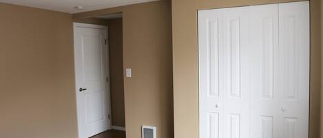 Una habitación con paredes color beige, una puerta blanca, una lámpara de techo blanca, una rejilla de ventilación y un cubo de basura.