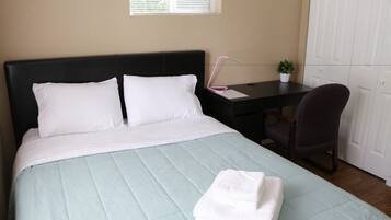 A neatly made bed with white bedding, a desk with a chair, a small potted plant, and a window with blinds.