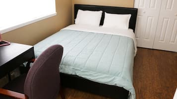 En pænt redt seng med et lyseblåt quilt, et træskrivebord med stol, og en hvid dør i baggrunden.