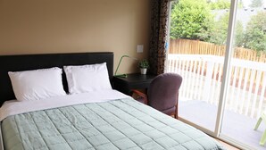 A bed with a green quilt, a black headboard, and white pillows. There is a small table with a plant and a chair. A window with a view of a wooden fence and greenery is visible.