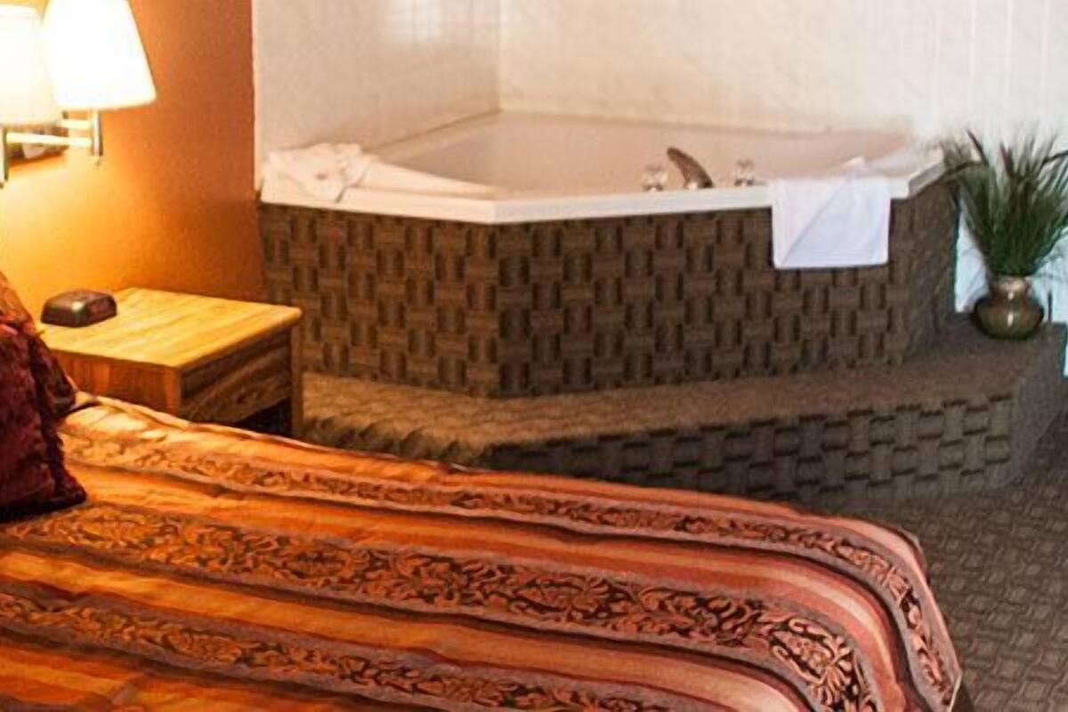 A hotel room with a bed, a nightstand, a lamp, a painting, a bathroom with a bathtub, and two potted plants.