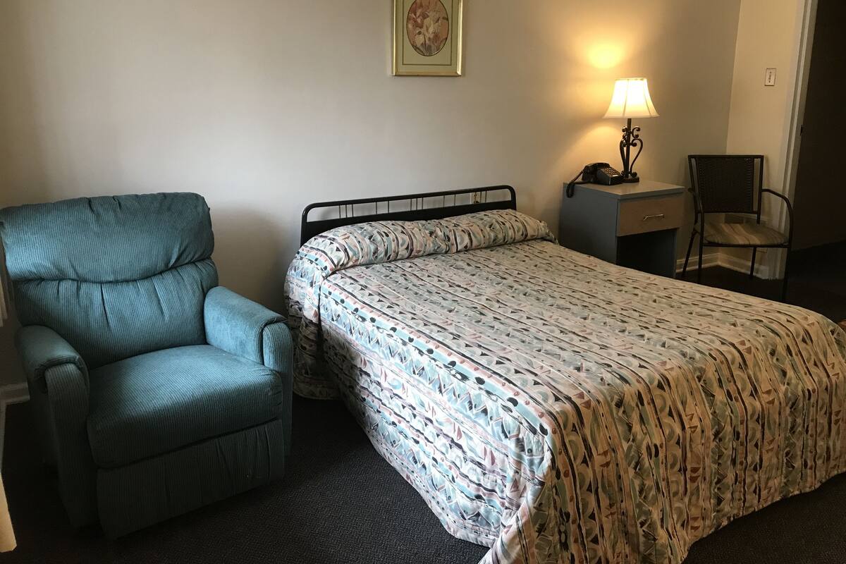 A room with a bed, a chair, a desk, a lamp, and a framed picture on the wall.