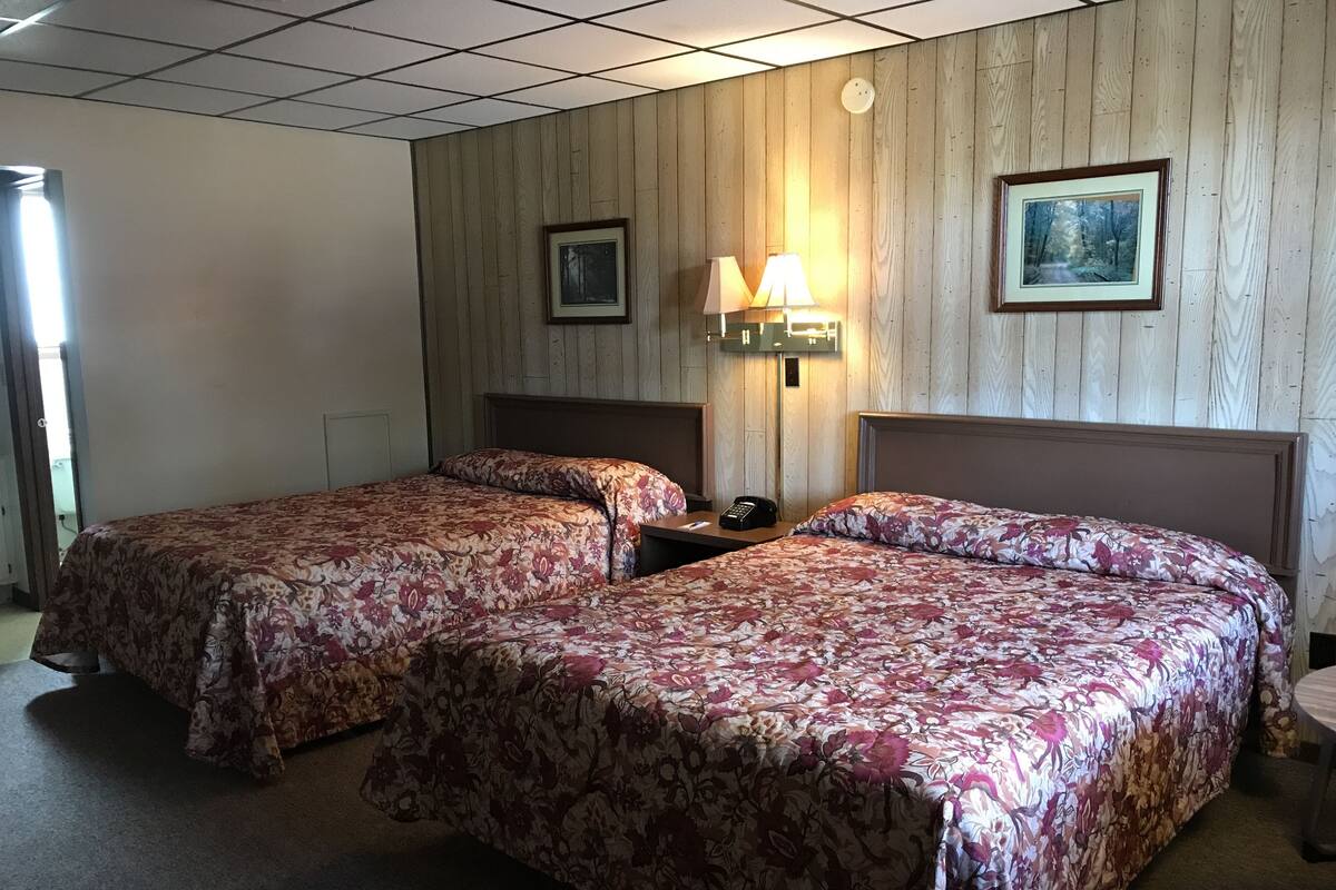 A hotel room with two beds, wooden panel walls, a nightstand with a lamp, and framed pictures on the wall.